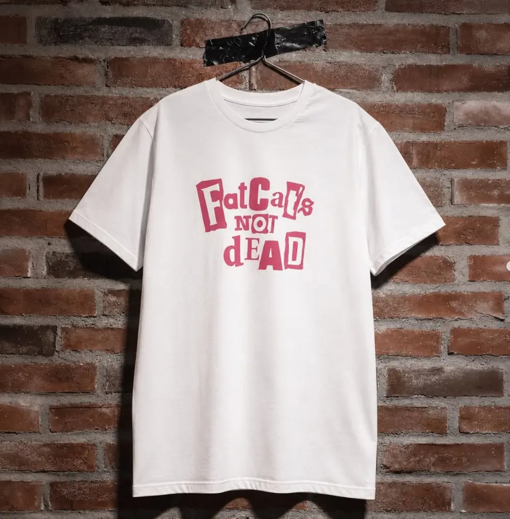 Weißes T-Shirt mit magentafarbenem Schriftzug Fat Cats Not Dead vor Backsteinwand